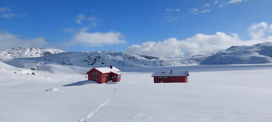 Tomannsbu. 2 røde hytter i snødekt landskap. Det er blå himmel og sol. 