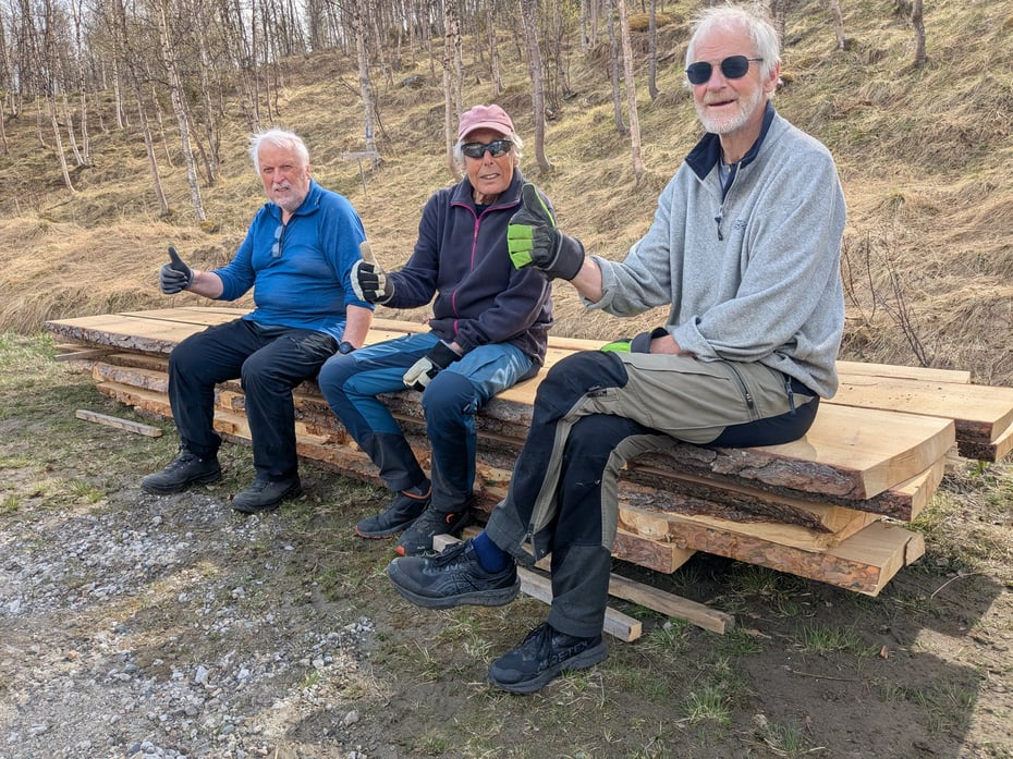 Tre eldre menn sitter på en stabel av kraftige planker. De viser tommel opp. 