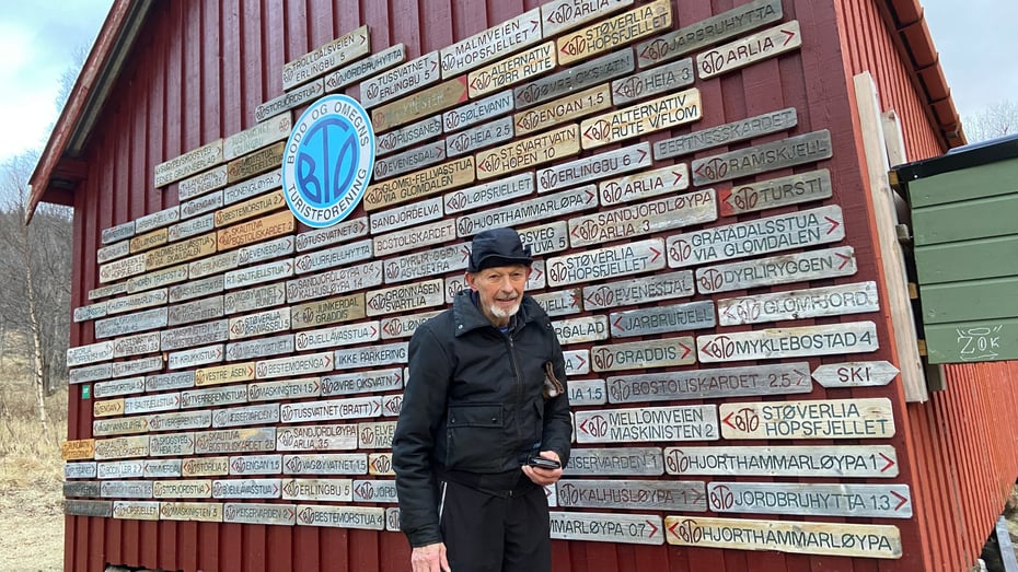 Eldre mann foran en rød vegg dekket med treskilt og BOT-logoen