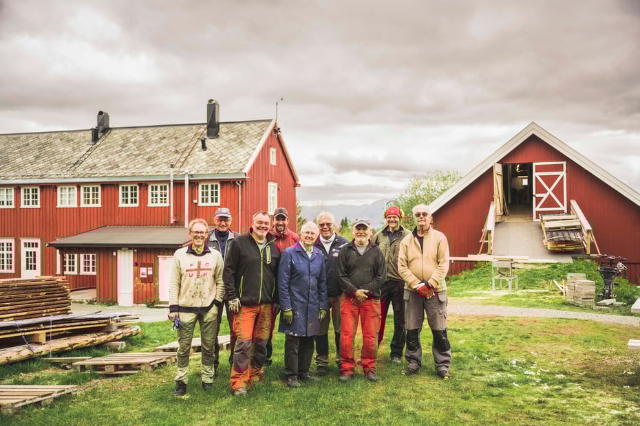 Ni personer stilt opp på to rader ute på et gårdstun, med en rød hytte og en rød låve, begge med hvite lister, synlige bak dem.