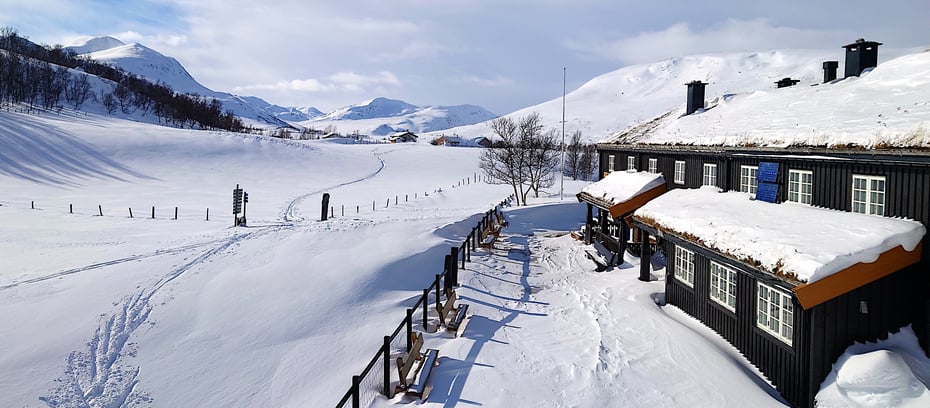 Langsiden av en brun to-estasjes hytte tar opp høyre kant av bildet, med et gjærde foran som skillet det lille tunet foran hytta fra det store og snødekte landskapet som strekker seg utover i resten av bildet. Helt hvit-dekte fjell i bakgrunnen. 