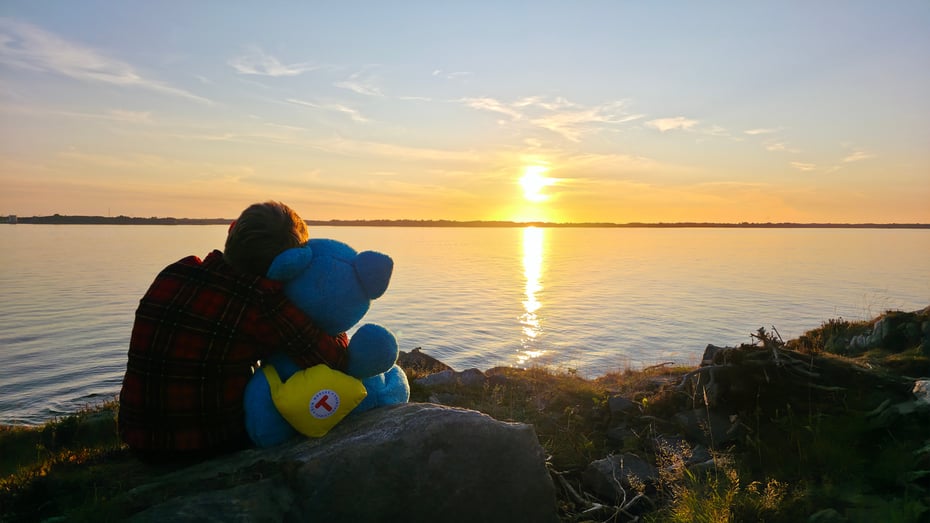 Gutt sitter foran solnedgang og holder rundt blå bamse.