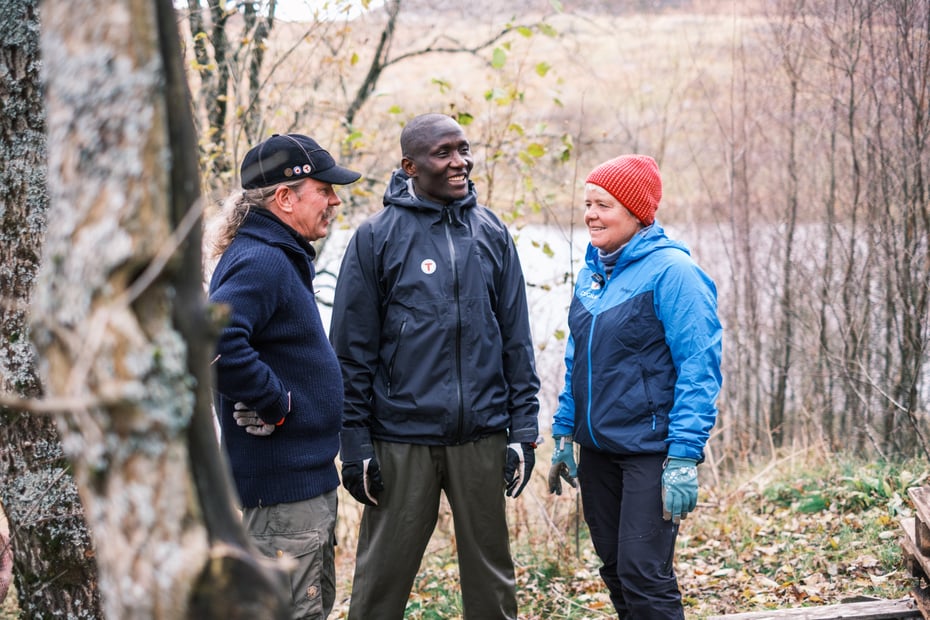 Tre personer står og samtaler i et skogsholdt, høstlige farger på naturen rundt. 