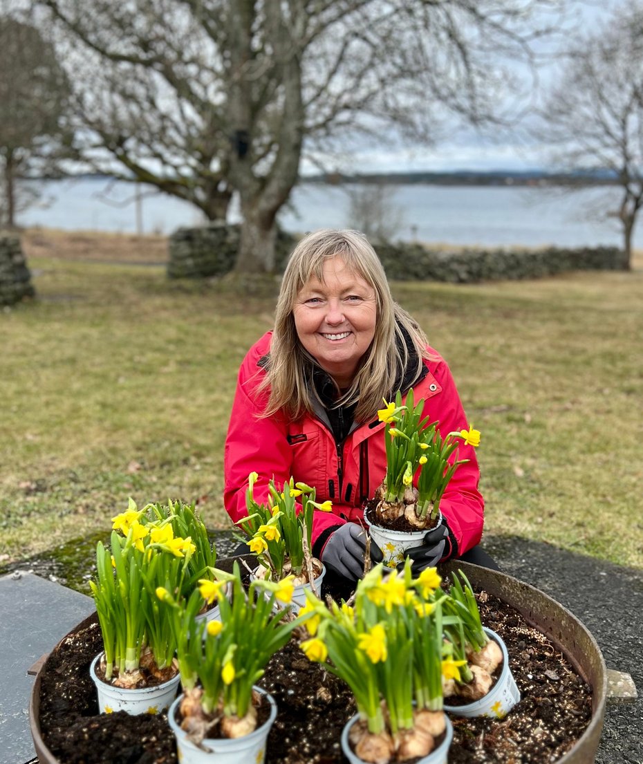 Kvinne i rød jakke sitter bak en potte med blomster
