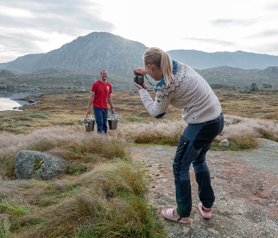 Kvinne i ullgenser fotograferer mann med rød t-skjorte som bærer to bøtter. De står i naturen