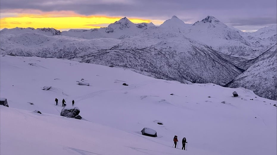 En gruppe mennesker som beveger seg gjennom et vinterfjell-landskap med soloppgang i bakgrunnen. Lyset langs horisonten er varmt – gult og oransje – og står i sterk kontrast til de kalde blå og hvite tonene i snøen og fjellene rundt.