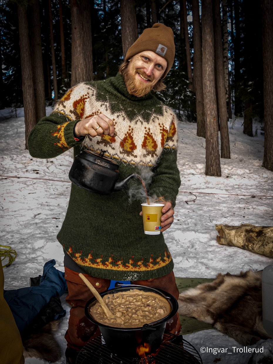 En smilende mann i skogen med en kaffekanne og kopp i hendene. På bålet står en gryte med mat. Foto.
