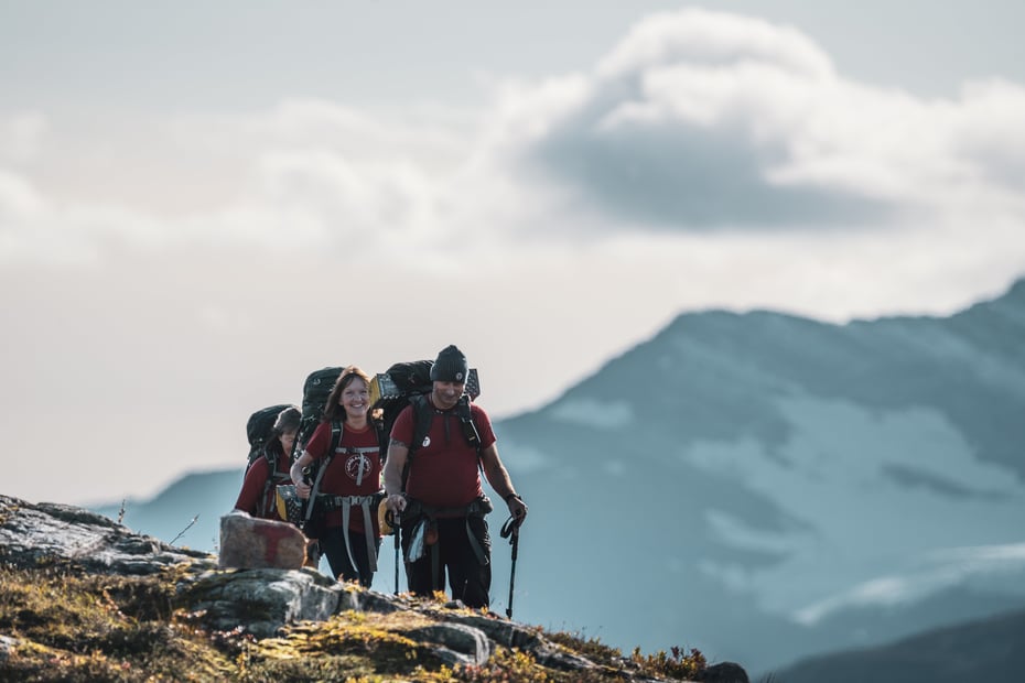 Tre mennesker med tursekker går i et fjellandskap. Et stort, snøkledt fjell i bakgrunnen. Alle går i T-skjorte, og mannen som går fremst har lue og gåstaver.