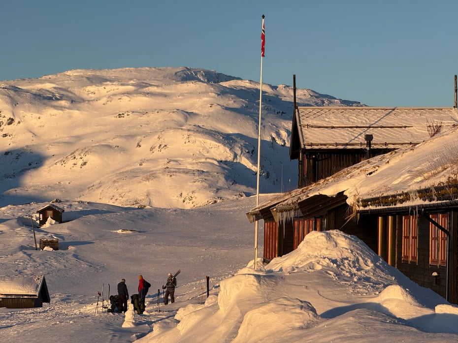 Foto. Haukeliseter fjellstue i ettermiddagslys. Snødekte fjell i bakgrunnen, tre personer står på tunet.