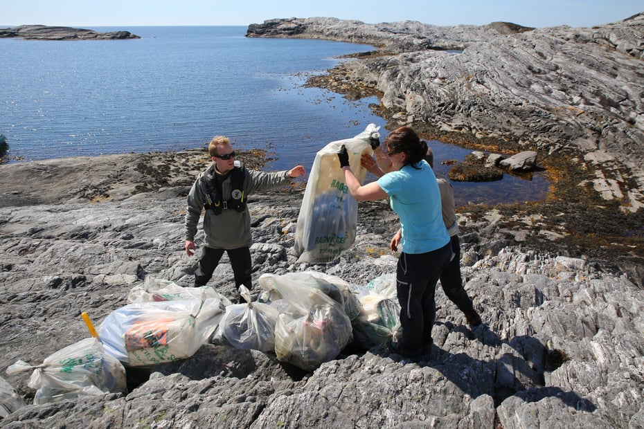 To mennesker rydder plast i strandsona en sommerdag med sol og blå himmel.