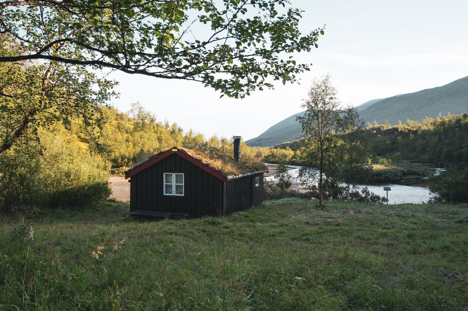 Ei mørkebrun hytte med torvtak. Foran er det grønt gress og en furukvist vises i venstre hjørne. Bak er det en elv og skoger.