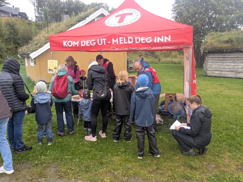 Samling av folk foran paviljong med DNT-logo. Gammelt gult hus i bakgrunnen