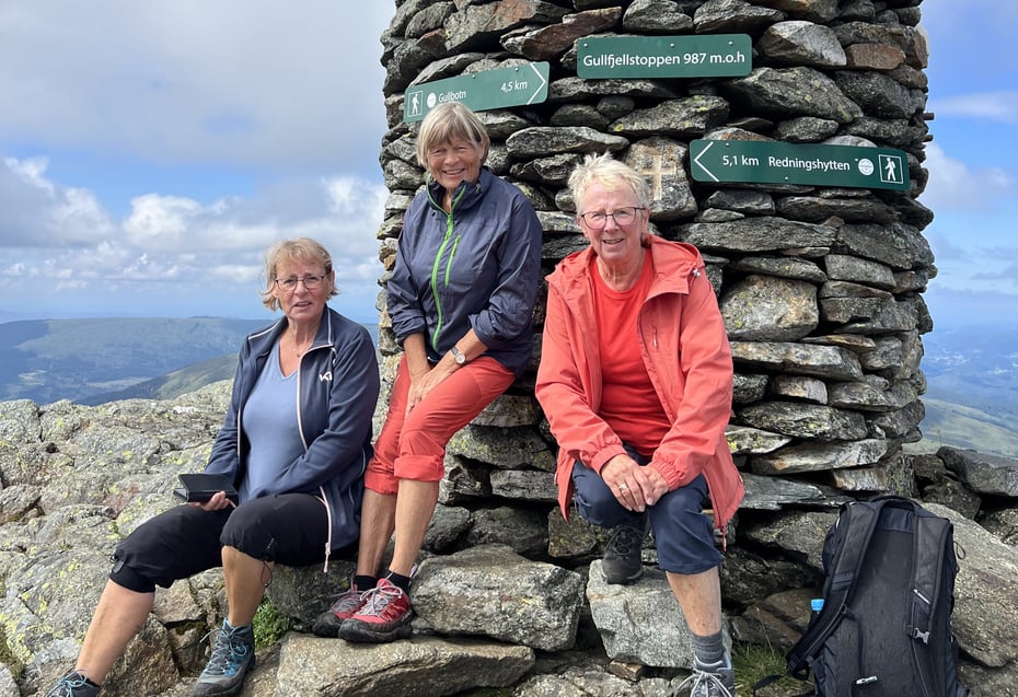 Tre damer sitter på fjelltopp foran varde