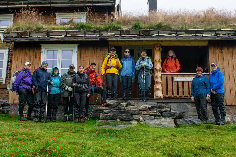Gruppe med svaksynte turgåere med ledsagere utenfor gammel tømmerhytte.