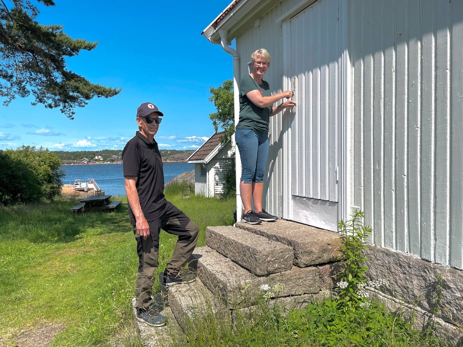 Mann og kvinne som står på trappen til et hvitt hus ved kysten. Foto.
