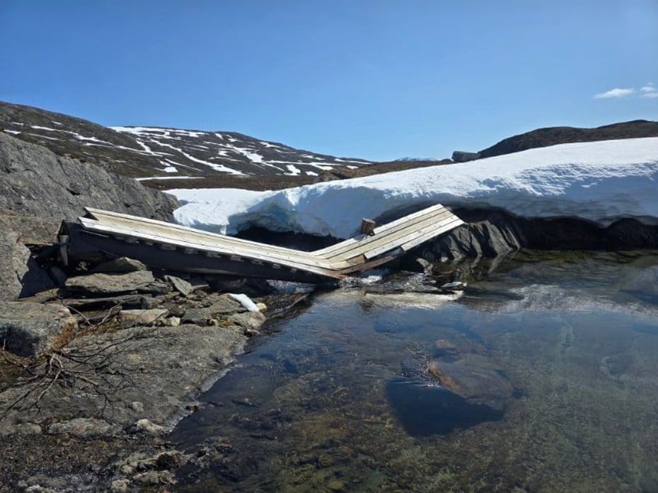En trebru som er brukket i midten. Foran er det vann, og bak er det mye is og snø. Det er et steinete landskap. 