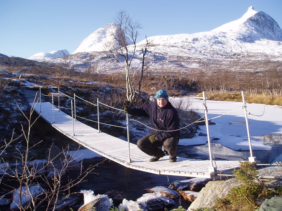 Bru med en person på og snøkledte fjelltopper i bakgrunnen