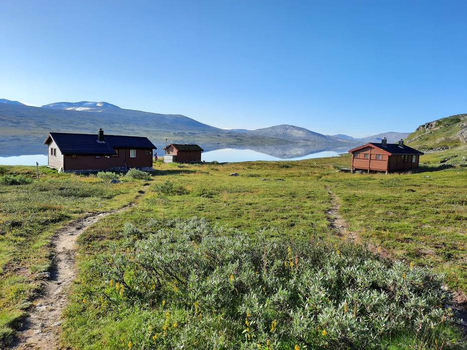 Tre brune bygninger på grønn bakke med blankt vann og fjellformasjon i bakgrunnen