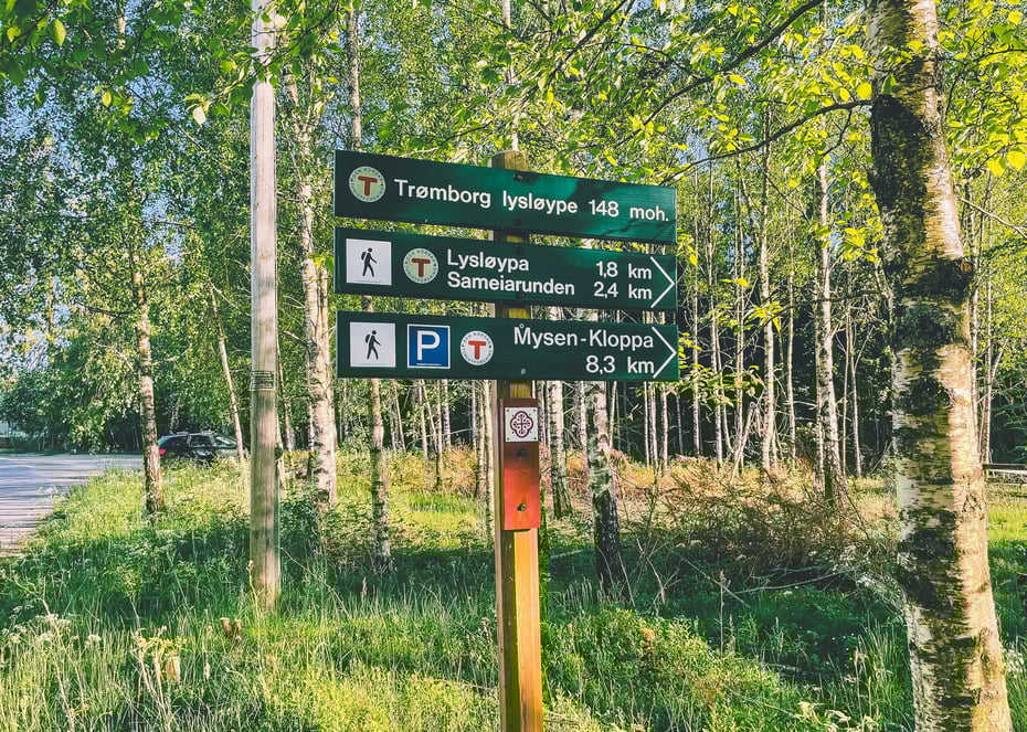 Stiskilt i et skogsområde viser vei og avstand til Trømborg lysløype, Lysløypa Sameiarunden og Mysen-Klopa, sammen med gang- og parkeringssymboler.
