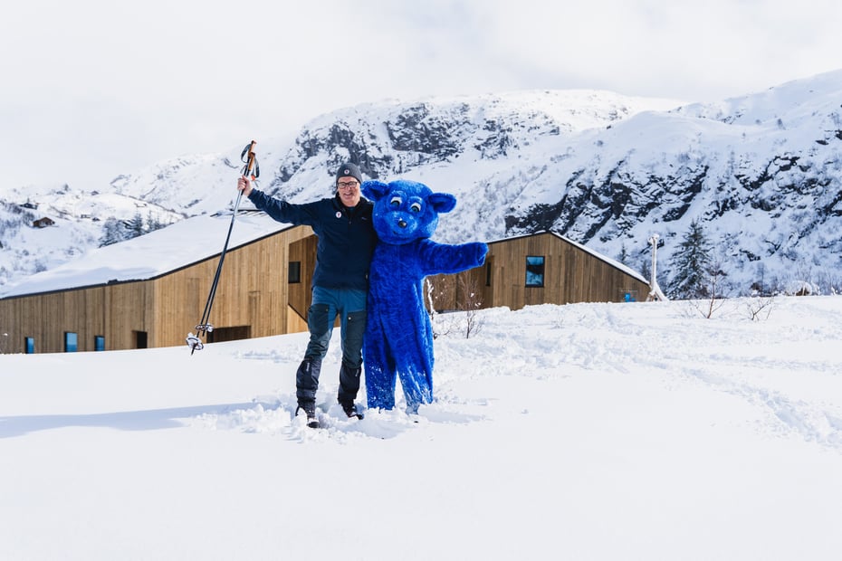 Mann og blå bamse står i snøen utenfor en hytte