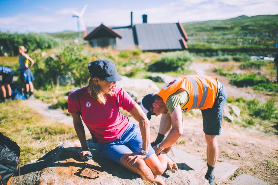 En mann med oransje refleksvest ser på hælen til en kvinne som sitter på en stein.