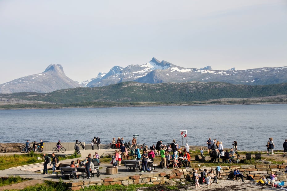 en stor gruppe mennesker feirer Sankthans ved sjøen. I bakgrunnen er det høye fjell med snø på.