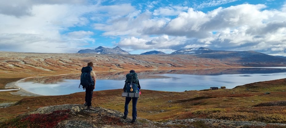 To gutter med tursekker står på en høyde og ser ut over et høyfjellandskap med topp og et digert vann foran. Det ligger noen hytter nede ved vannet. 