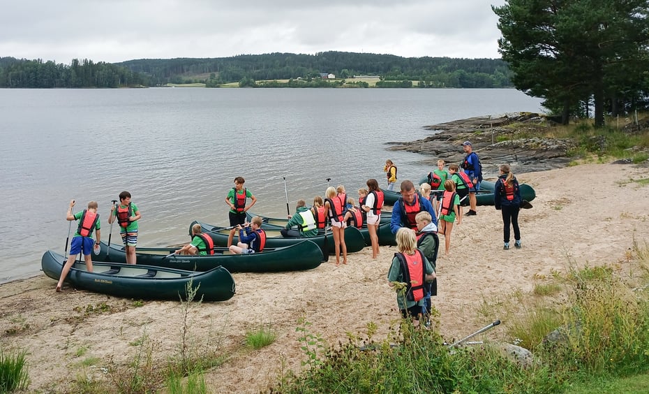 En gruppe barn/ungdommer setter på seg flytevester og klargjør kanoer for en padletur på en liten strand.