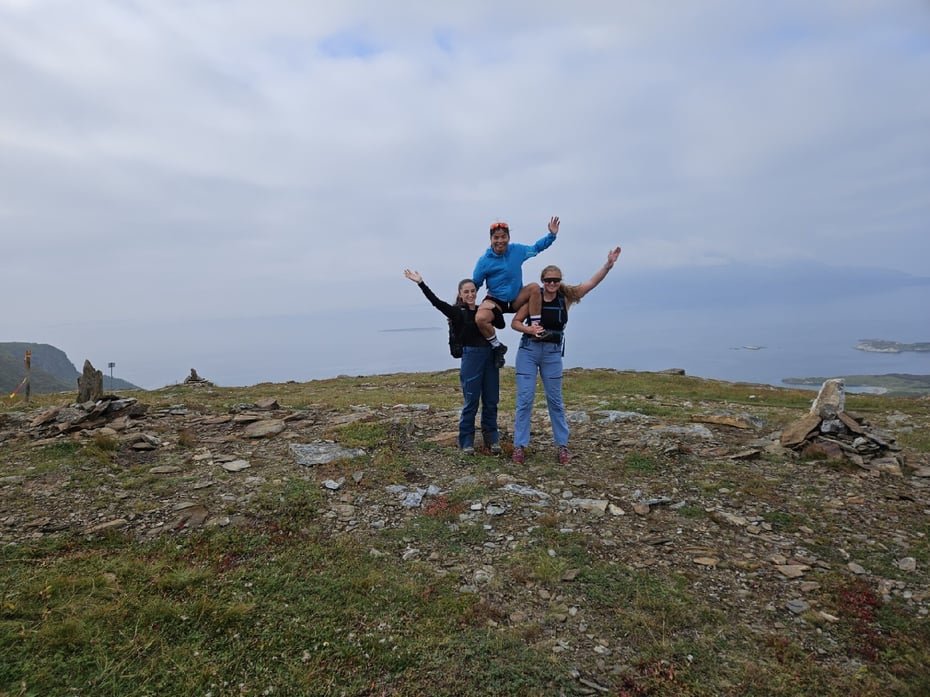 Tre ungdommer står på toppen av et fjell. En av dem sitter på skuldrene til de to andre. De holder armene i været og smiler. 