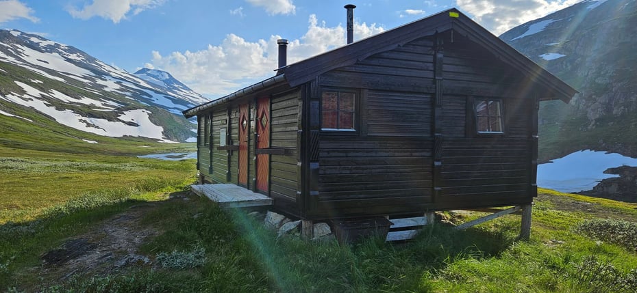 Ei brunmalt hytte i høyfjellandskap. Det er snøkledte fjelltopper i bakgrunnen. Sola skinner og det er lettskyet.