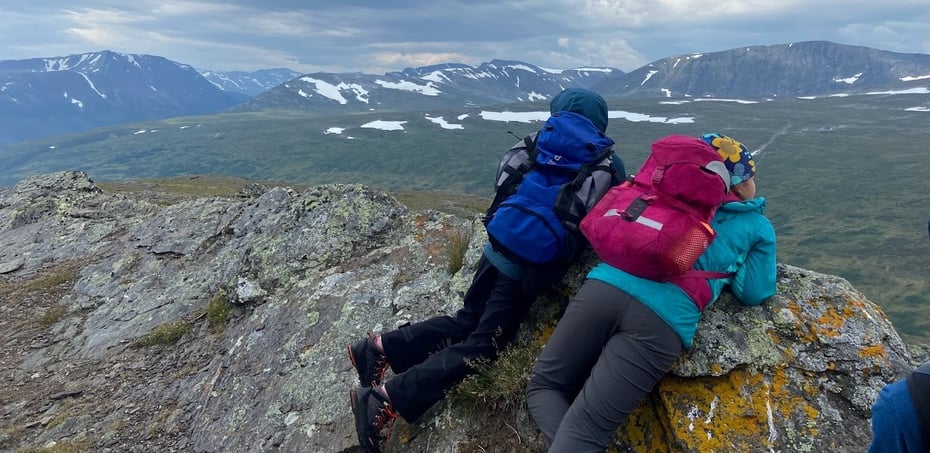 To barn i turklær og fargerike tursekket på ryggen, ligger på magen på en fjelltopp og ser utover det store landskapet som strekker seg ut under dem. Fjellkjede kan sees langs horisonten, under en grå og overskyet himmel. 
