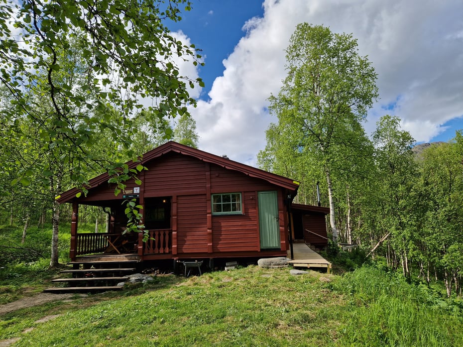 Ei rødbrun hytte med bjørketrær rundt. Blå himmel med hvite skyer.