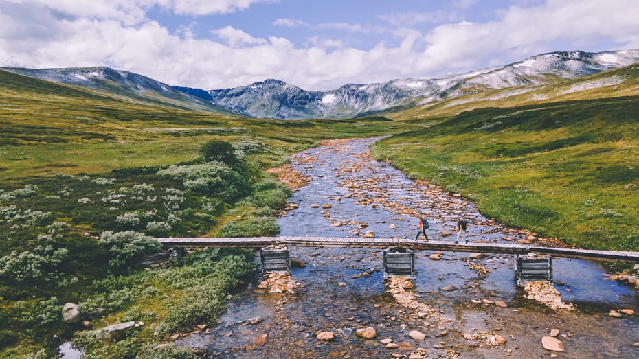 En smal bru over en vid og grunn elv ute i naturen, med grønne sletter på hver sin side. To personer er en tredjedel over brua.