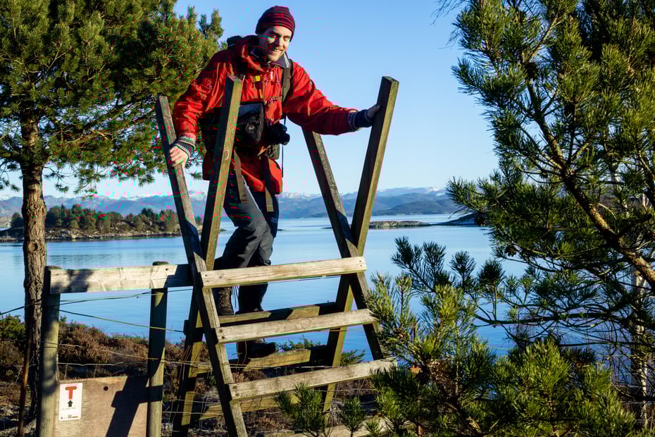 Mann i rød anorakk klatrer over gjerdeklyver med fjord i bakgrunnen.