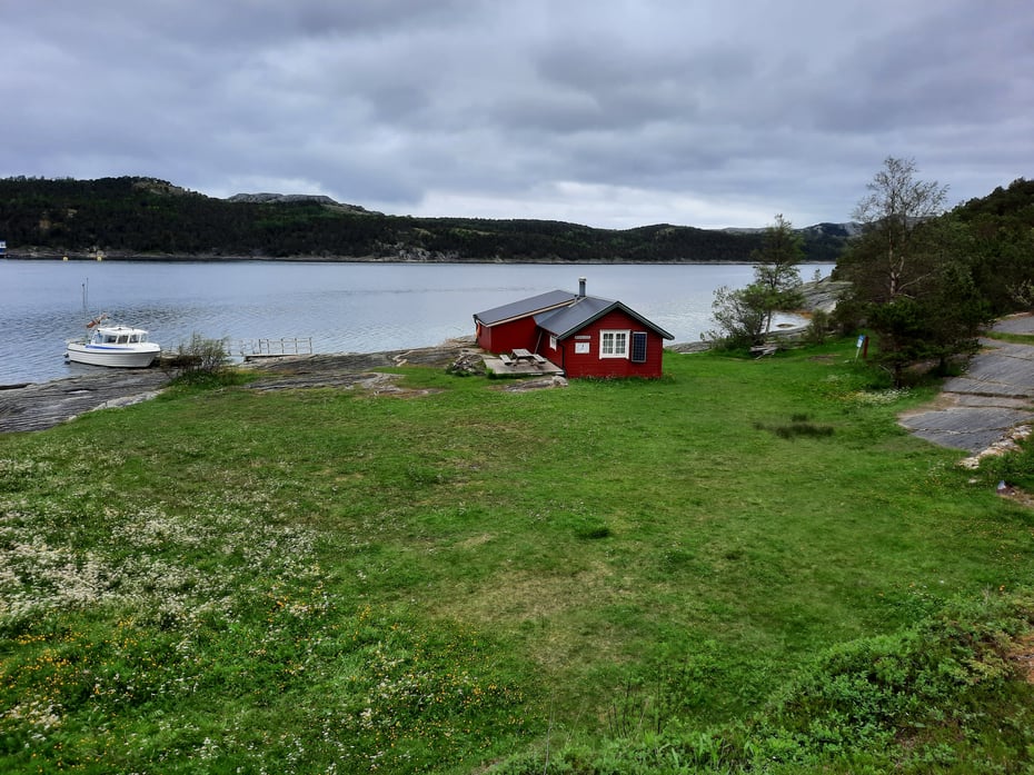 Egnarbua er Norges lavestliggende DNT-hytte. Bilde av rød hytte med gress foran og sjø og fjell bak. En hvit båt til venstre i bildet. 