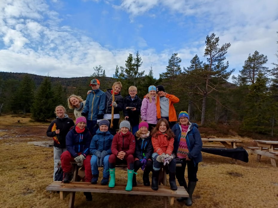 Barn og voksne sitter på en benk og smiler til kamera, bak er det skog. 