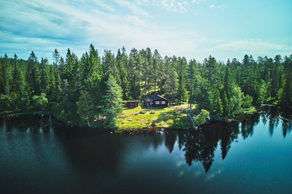 En koselig hytte ved vannkanten omgitt av tett skog på en klar og solfylt dag.
