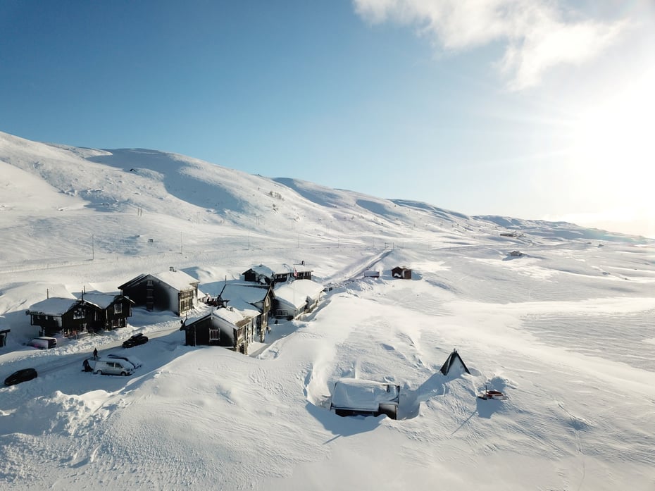 Oversiktsbilde av Haukeliseter fjellstue fra februar 2023, bygningende er dekket av snø og sola skinner over et snøkledd landskap.