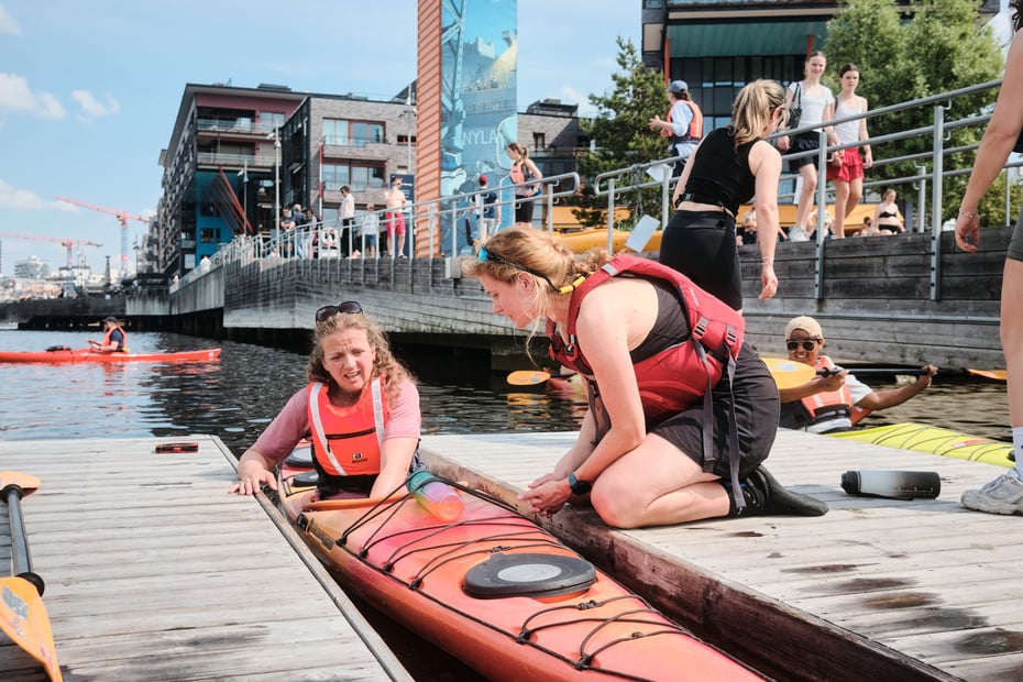 En person som sitter på en brygge og hjelper en annen person i en kajakk
