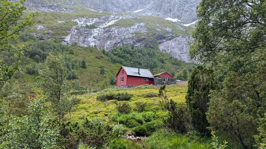 Liten rødmalt hytte ligger på en bø mellom fjell og trær.