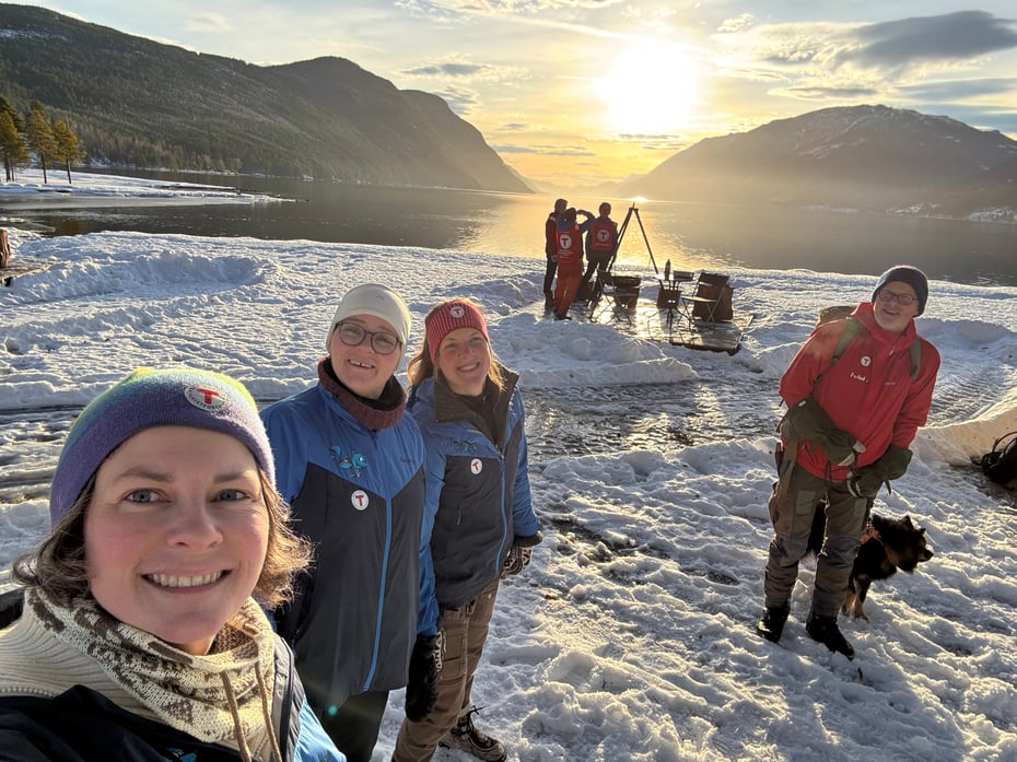 Smilende mennesker som står på en snødekt strand med et bål og fjell i bakgrunnen.
