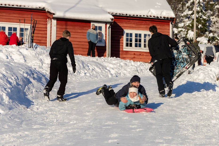 To jenter som smiler på akebrett ned bakke med rød huyttevegg bak dem.