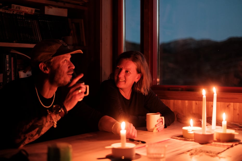 To personer sitter ved et bord med hver sin kaffekopp. I bakgrunnen blåtimen over fjell. Stearinlys brenner på bordet.