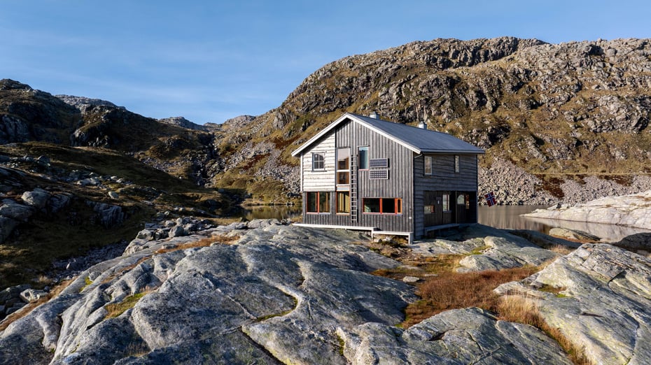 Stor, grå hytte med store vinduer og skråe vinkler i solrikt høstfjell.