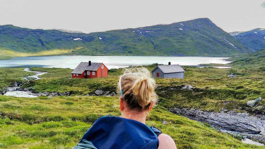 Kvinne med lyst hår og med ryggen til ser mot en rødmalt hytte i det fjerne.