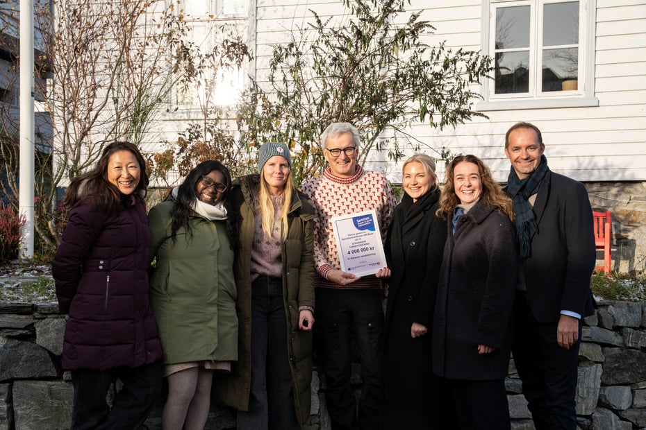 Syv personer står foran Stavanger Turistforening sitt kontorlokale og holder frem en gavesjekk