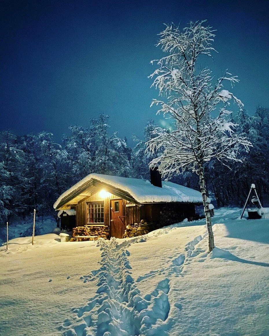 Ei tømmerhytte med snø på taket i et snøkledt landskap. Det er mange bjørketrær i bakgrunnen. Det er skumring, og et lys står på over inngangen til hytta.