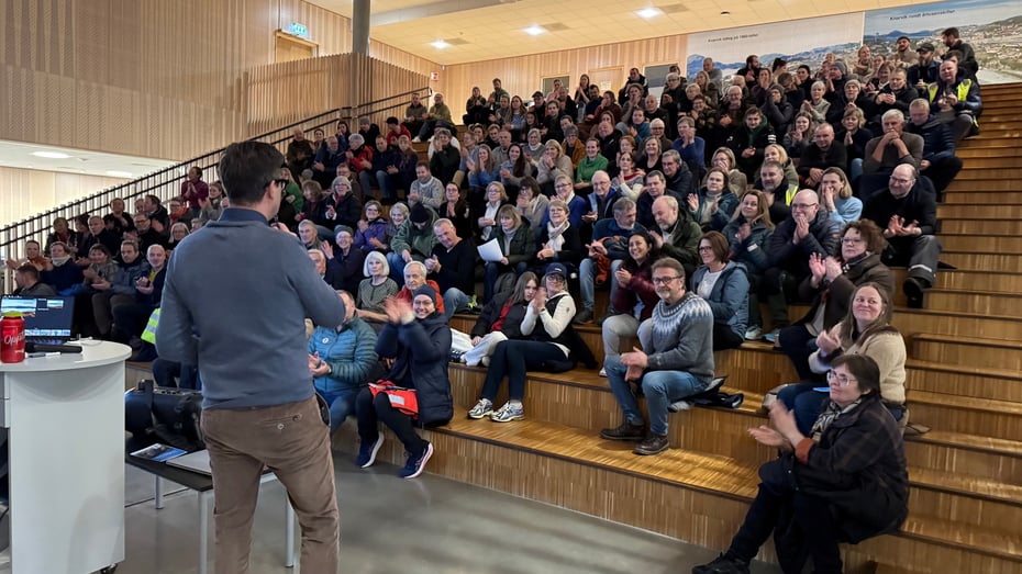 En mann står foran en stor forsamling med folk som smiler og klapper.