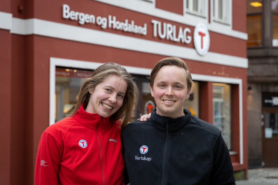 Kvinne i rød genser og mann i blå genser smiler til kamera for Turlagets butikk i Bergen.