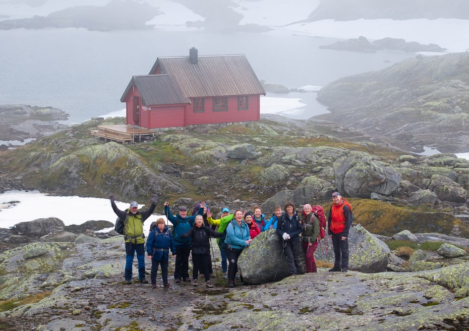 En glad gjeng smiler foran en rødmalt hytte i fjellet med tåke rundt hytten.